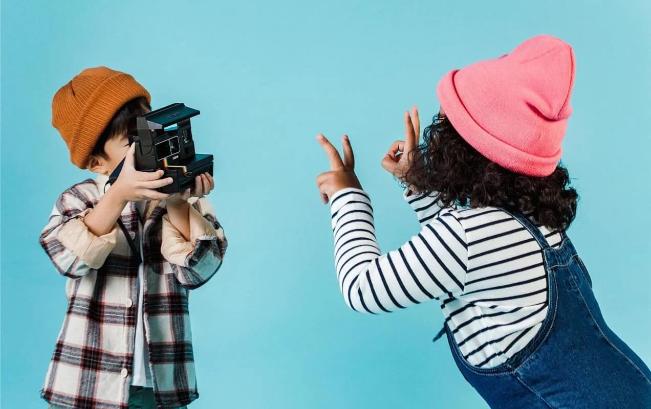 boy in a plaid shirt using an old-fashioned camera to take a photo of a girl in overalls and a striped shirt