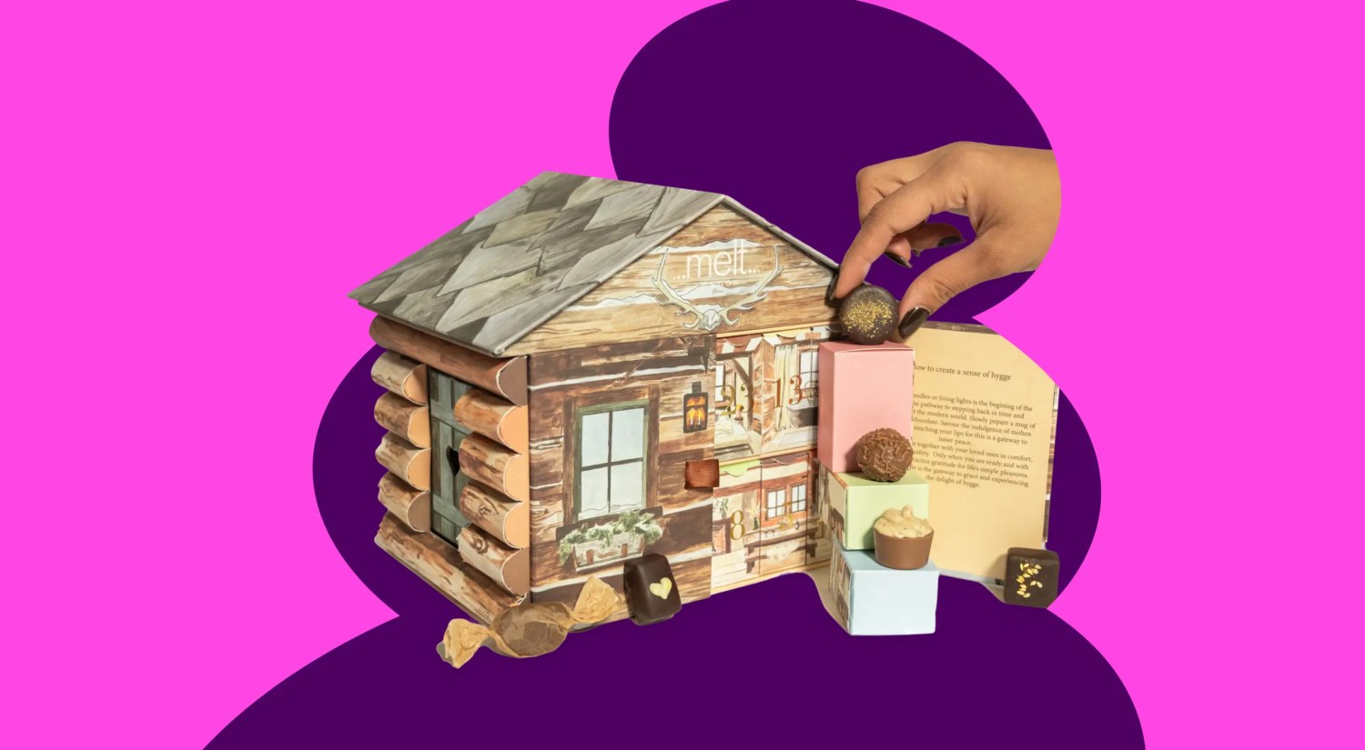 a photo of a miniature log cabin advent calendar with three chocolates on display, on a dark and light pink background