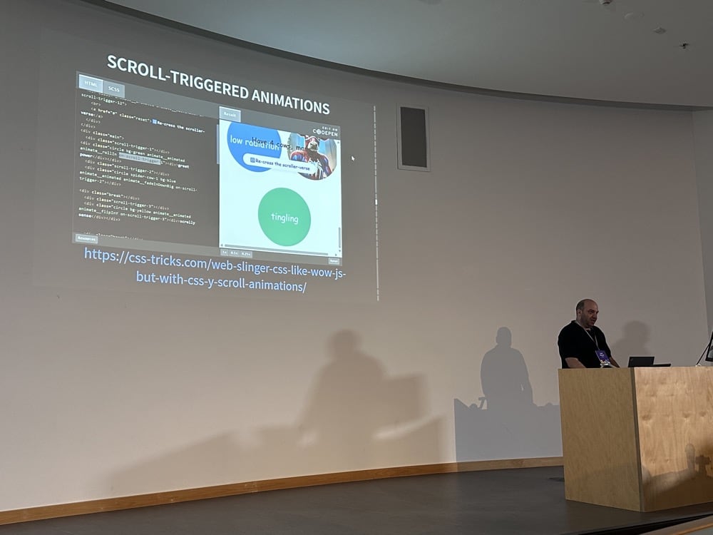 Lee Meyer speaking at light wooden lectern in front of a projection showing a demo of his scrollytelling library on CodePen.
