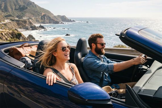 Male and female in a convertible auto driving alongside the ocean
